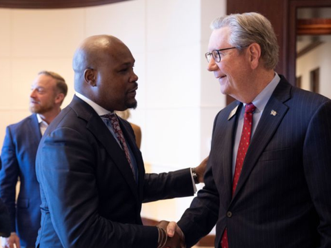 Two men in suits shake hands and smile at each other in a formal indoor setting. Other people in business attire are visible in the background.
