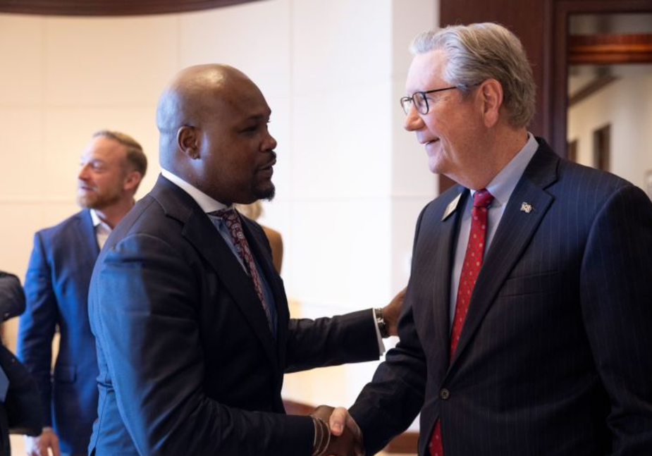 Two men in suits shake hands and smile at each other in a formal indoor setting. Other people in business attire are visible in the background.