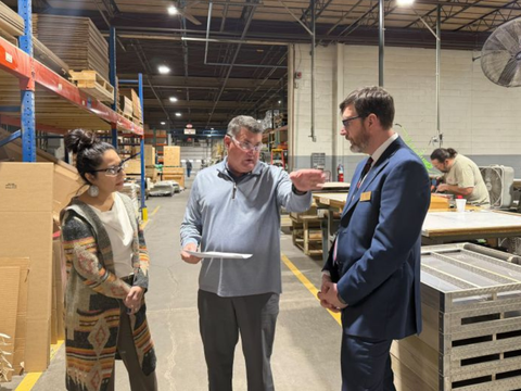 Three people stand in discussion inside a warehouse; one man gestures while holding papers, speaking to a woman and a man in a suit. Shelves with boxes and workers at tables are visible in the background.