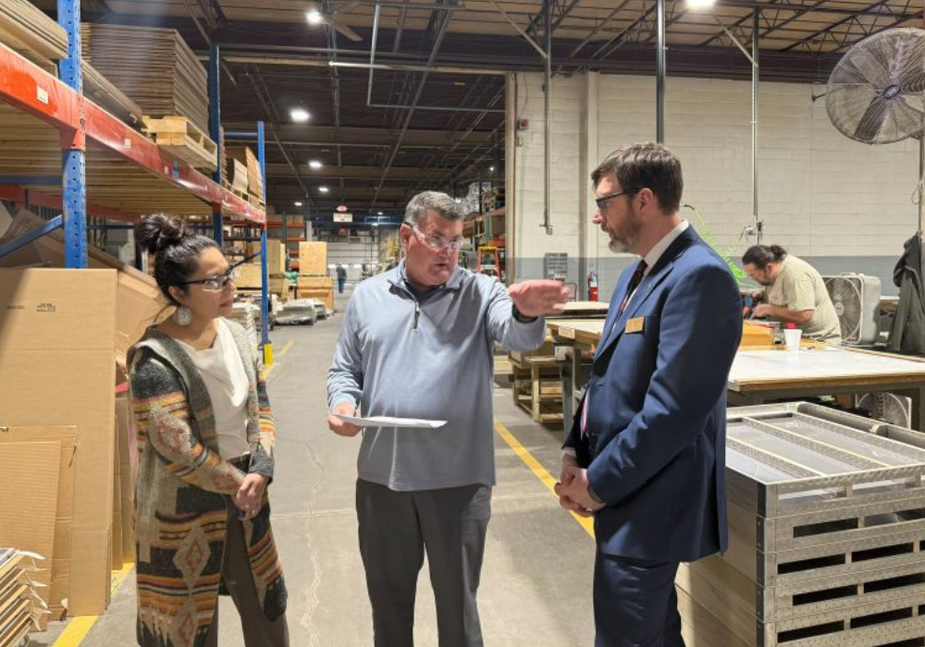 Three people stand in discussion inside a warehouse; one man gestures while holding papers, speaking to a woman and a man in a suit. Shelves with boxes and workers at tables are visible in the background.