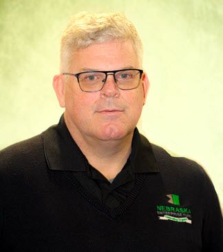 The photo shows a white man in a black polo shirt with the logo of the Nebraska Enterprise Fund on the front. The main has blonde hair and is wearing glasses. He appears to be in his mid-40s.