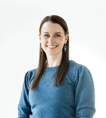 A White woman in her 20s with long dark hair, wearing a blue sweater, is smiling at the camera.