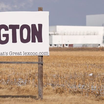 A large sign in a dry cornfield reads “Welcome To LEXINGTON a better life See What’s Great lexcoc.com.” In the background are large white industrial buildings under a pale sky.