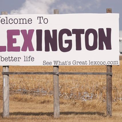 A large sign in a dry cornfield reads “Welcome To LEXINGTON a better life See What’s Great lexcoc.com.” In the background are large white industrial buildings under a pale sky.