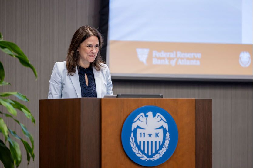 A woman with shoulder-length brown hair, wearing a gray jacket, stands at a podium.
