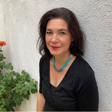 Photo of Monica Abeita, a black-haired woman in a black dress leaning against a white stucco building.