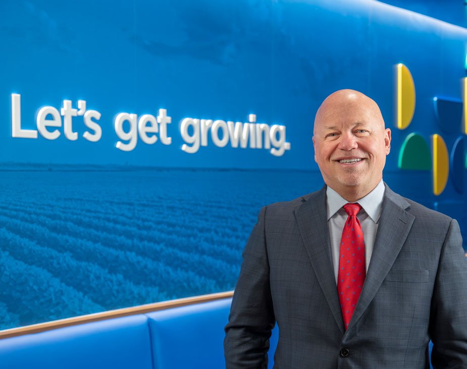 Paul Maass of the Kansas City Fed's Omaha Branch Board of Directors standing in a suit in front of a sign saying "Let's get growing" at his place of work, Scoular.