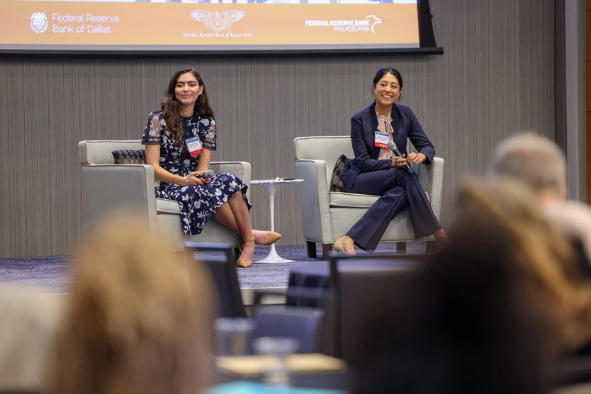 Two smiling, dark-haired women in their 30s sit on gray chairs on a stage in front of an audience.