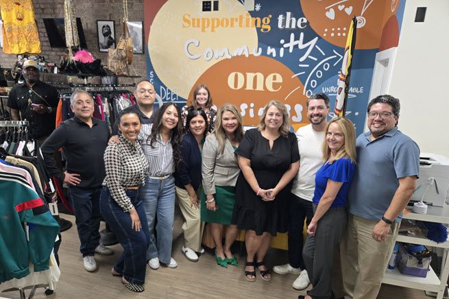A group of eleven smiling humans - seven women and four men - stand in front of a colorful mural painting on the wall inside a local coffee shop.