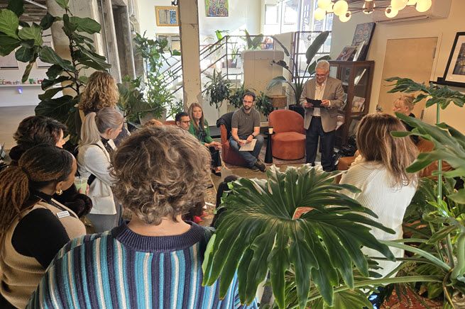 A group of people gathers at a local coffee shop. They're all facing a white-haired, smiling man, who is presenting information. The group is surrounded by potted plants.