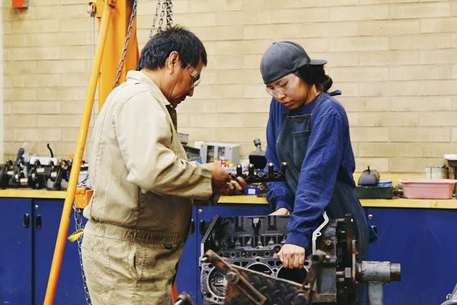 A Navajo woman takes an automotive class at her high school. The Navajo man who is instructing her helps her use a big metal machine.