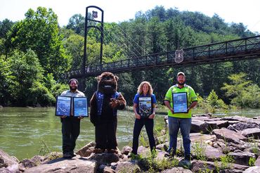 Two men, a woman, and someone in a bear costume hold up posters in front of a mountain stream.
