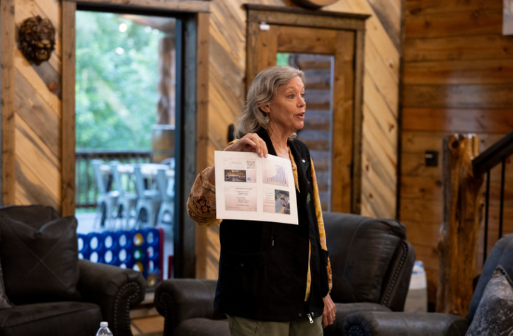 A woman stands in a cozy, wood-paneled room, holding up a paper with charts and images, as if presenting to a group. A window and outdoor patio with chairs are visible in the background.