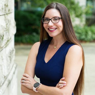 A White woman in her 30s has brown hair to her waist and is wearing glasses and a sleeveless navy blue shirt. Her arms are crossed in front of her and she's smiling at the camera.