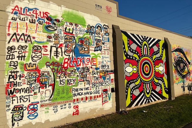 The photo is of a long wall in northeast Oklahoma City, a historically Black community. The wall looks like it's been turned over to artists. Three artists with different styles have tackled big squares of the wall. The first square is in graffiti style, the second is mod style in black, white, red and lime green, and the third is a giant eye, staring at you and not in a friendly way.