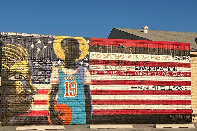 The photo is of a mural in northeast Oklahoma City. It's wall-sized and rectangular. Imagine a big flag, with a tall young Black man holding a basketball breaking up the space between the field of stars and the stripes. On the strips is written a quote by Ralph Ellison, some of which is illegible. The last line are, "on this day of emancipation...let's us tell ourselves our story."