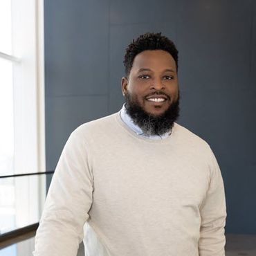 African-American man with a beard, wearing a white sweater over a light blue shirt and standing in front of what appears to be a chalkboard.