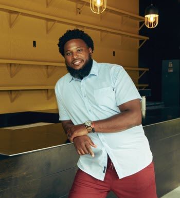 Photo of Quintin Hughes, a young Black man with a full beard, wearing a light blue shirt and maroon pants, leaning on a steel counter in a commercial space.