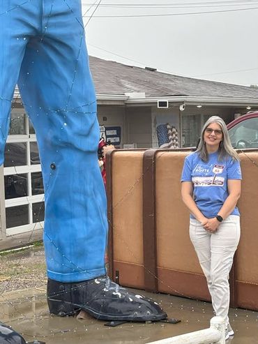 A smiling blonde woman stands by a 22-foot-tall fiberglass giant outside the diner she owns. She comes up barely above the giant's knee.