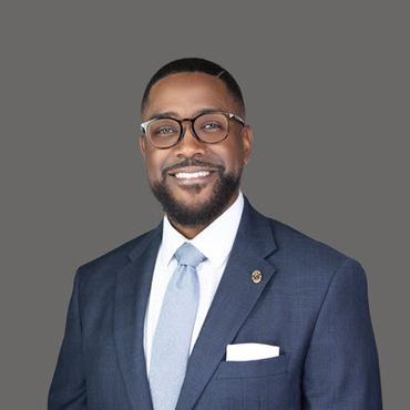 A Black man in his 40s with a short beard and short hair wears glasses, a navy blue jacket, a bright white shirt and a pale blue tie. He is smiling.