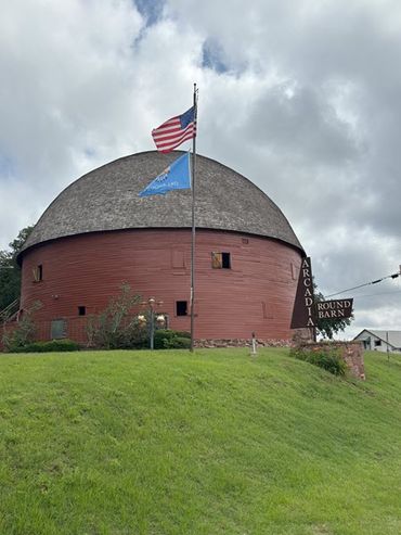 A round red barn with a domed roof stands at the top of a hill, with storm clouds behind it.