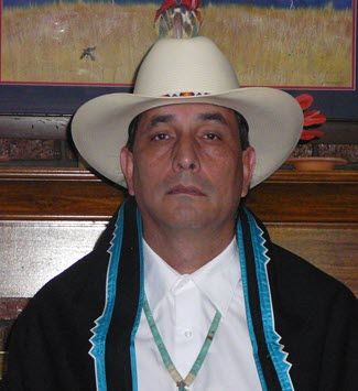 The photo shows a serious-looking Native American man in his 50s wearing a bright white shirt, turquoise necklace and white cowboy hat.