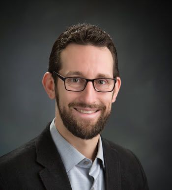 The photo is of a man in his 40s, smiling, wearing a black suit and blue shirt. He has eyeglasses and a beard.