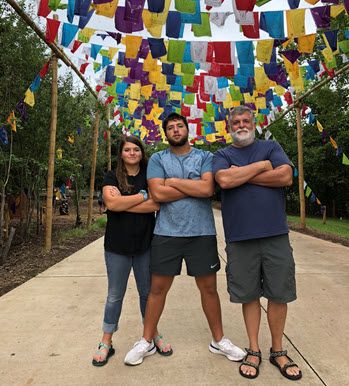 A young woman and young man with dark hair stand next to a man in his 50s with white hair and beard. They are all standing with their arms crossed, but they look amused rather than defiant. They're standing under rows of colorful banners, outside at a park or zoo.
