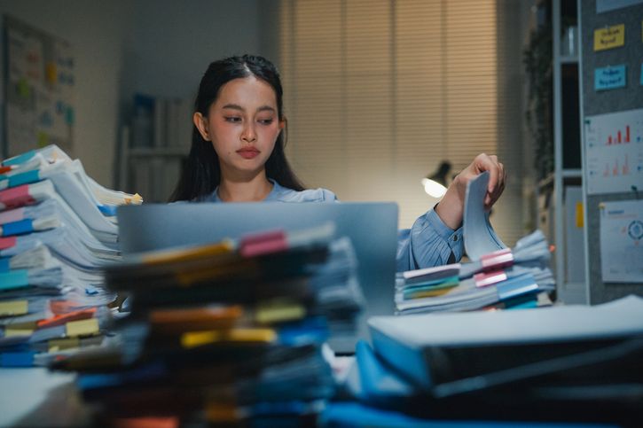 A young woman sits at her desk, surrounded by piles of folders with papers falling out.