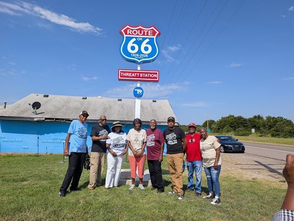 Seven members of the family, ranging in age from 40s to 70s, smile and stand together in front of a new sign marking their service station as a Route 66 monument.