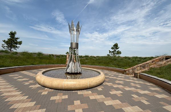 A fountain contains tall silver arrows in a low stone pond on a patio of brown and beige bricks. It's a panoramic view that shows trees and sky.