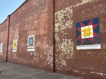 A long brick building, clearly old by the state of the bricks and the paint, has colorful barn quilts hanging down the side.