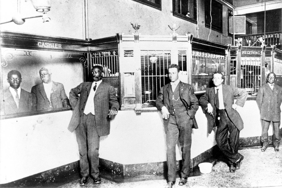 Image Description. Photograph of the interior of the Alabama Penny Savings Bank featuring six men of color wearing suits, standing next to the cashier window.