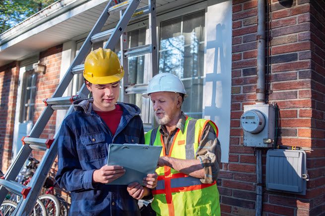 A young man and an older man examine a document. They are standing outside of a residence and there is a ladder in the background.