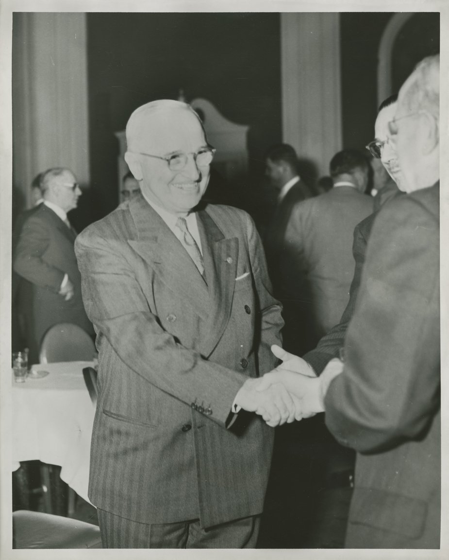 Image Description. Black and white photograph of President Truman smiling, shaking the hand of an unknown man in a large room with other men standing around tables.