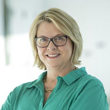 A White woman in her 50s with chin-length blonde hair is wearing a light green shirt and glasses. She is smiling.