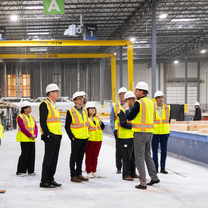 A group of people wearing hard hats and safety vests tour a large, well-lit industrial warehouse with construction materials and workers in the background.