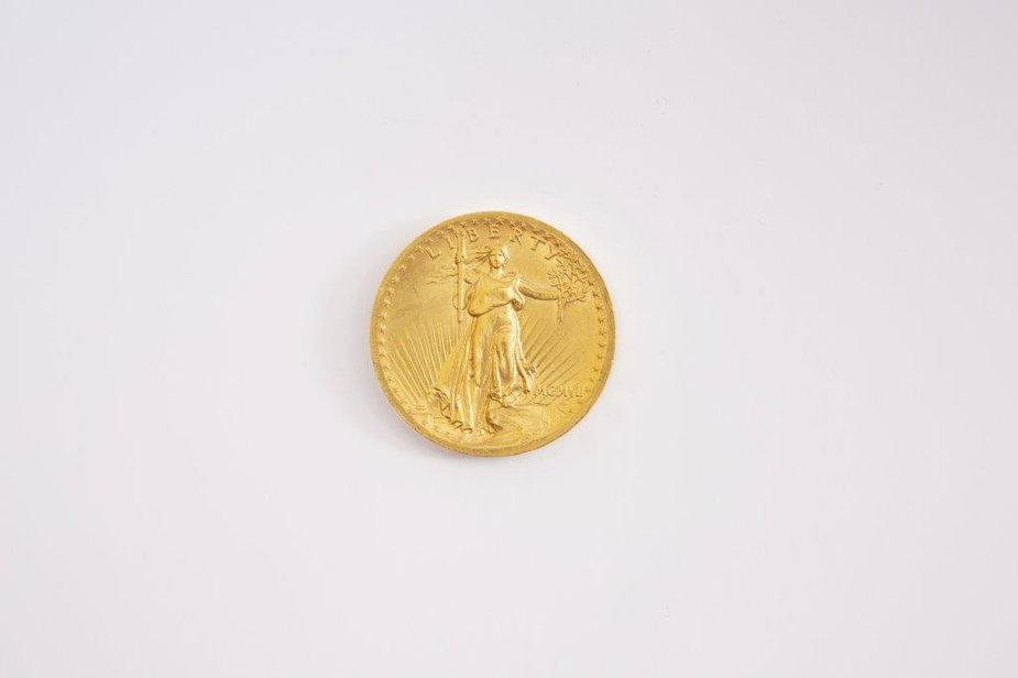 Image Description. The photograph shows a bright gold coin on a white background. The coin has an image of the model Hettie Anderson dressed as Liberty, holding a torch and an olive branch. The coin says "liberty" on it.