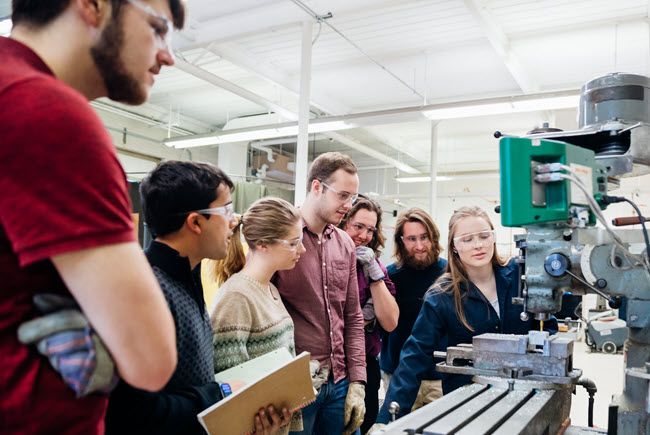 Six engineering students - a mix of male and female - gather around a big industrial machine while the instructor shows how to use it.