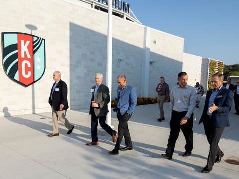 A group of six men in business attire walk and talk outside a white building with a large KC logo, under a clear blue sky.