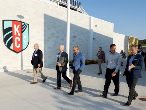 A group of six men in business attire walk and talk outside a white building with a large KC logo, under a clear blue sky.