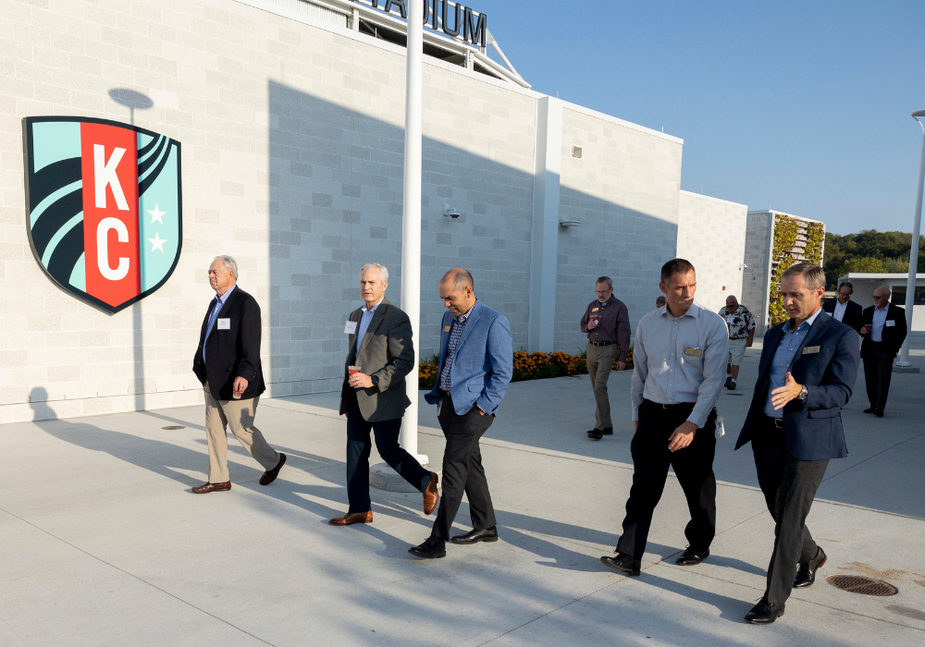 A group of six men in business attire walk and talk outside a white building with a large KC logo, under a clear blue sky.