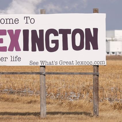 A large sign reads Welcome to LEXINGTON a better life in a brown field with an industrial white building in the background under a cloudy sky.