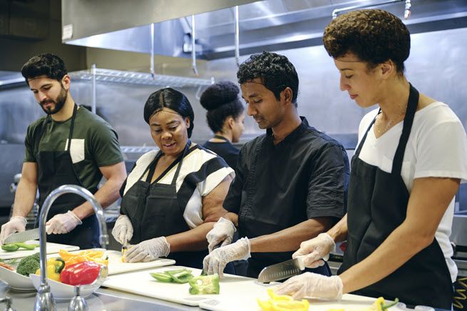 In an industrial kitchen, a female chef helps three male students perfect their knife skills by chopping vegetables.